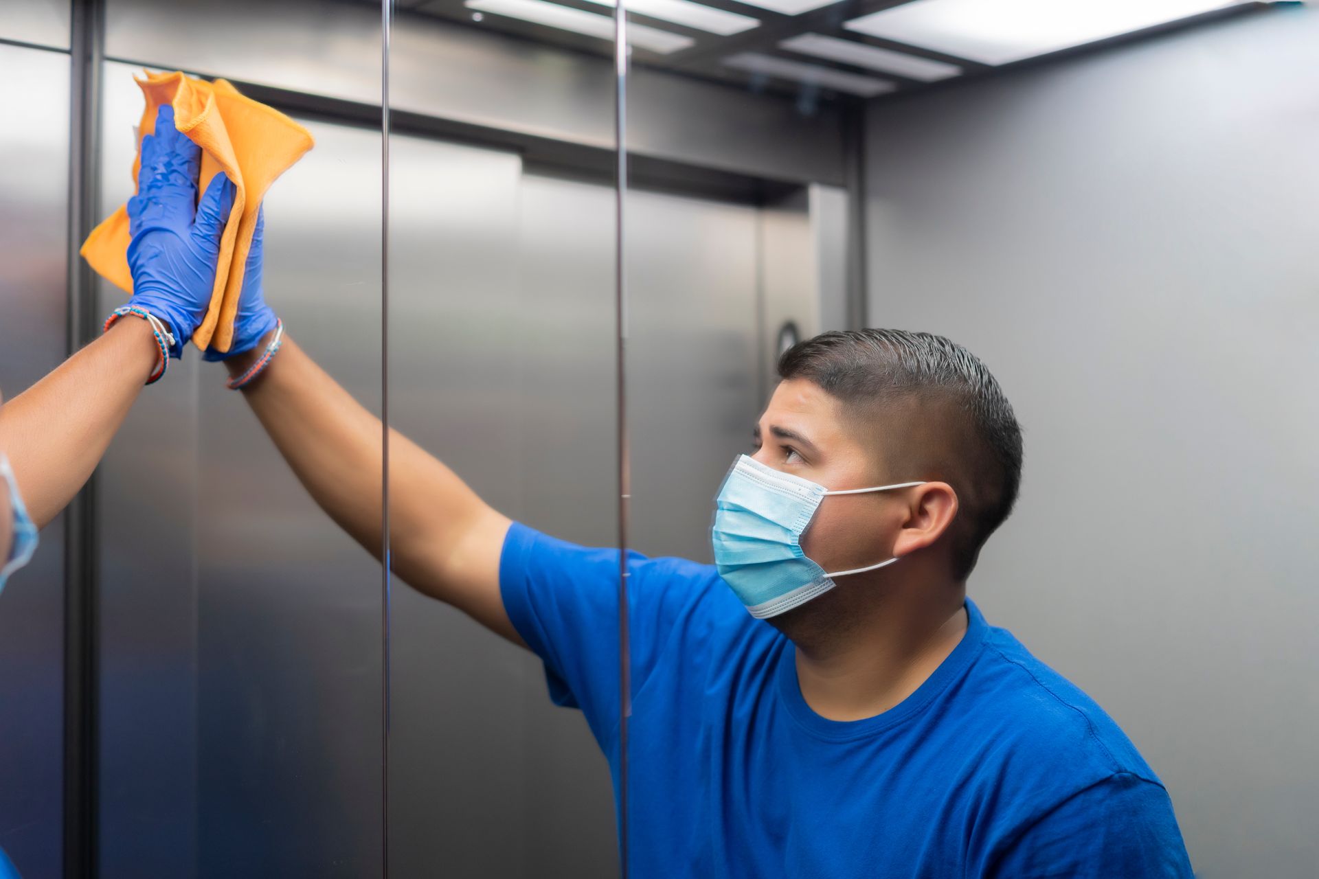 Masked cleaner wiping an elevator wall with a cloth while standing inside the elevator Masked cleaner wiping an elevator wall with a cloth while standing inside the elevator