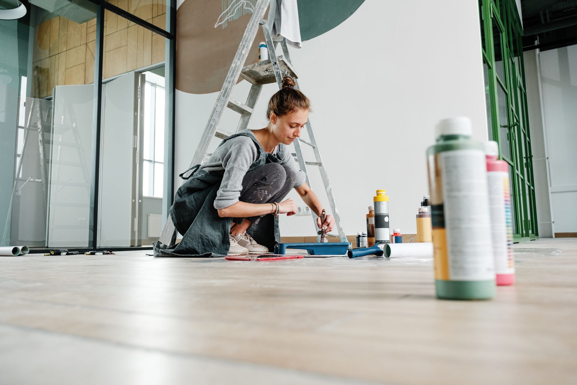 Person crouching on a wooden floor, painting or repairing a wall in a bright room with tools and ladders nearby