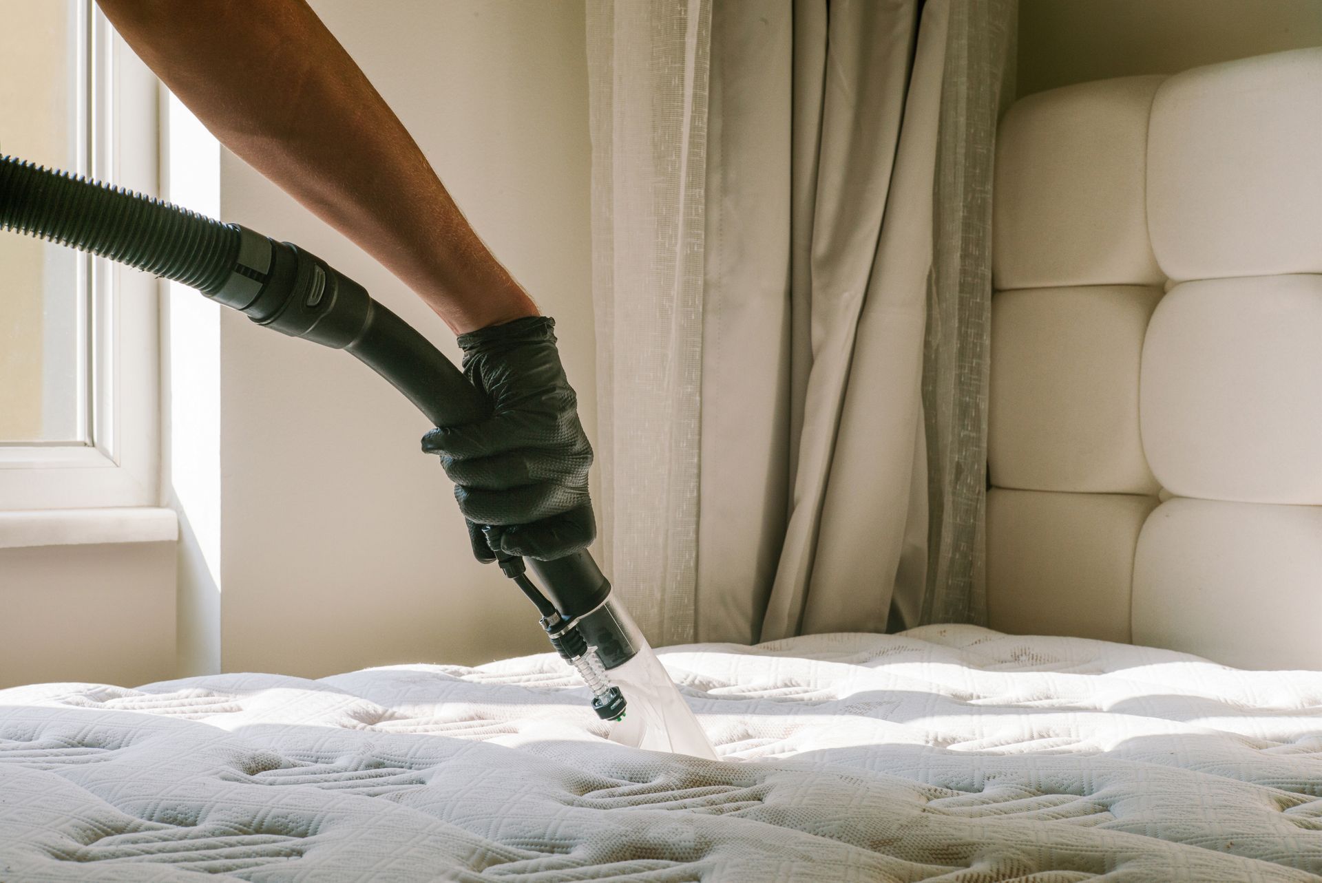 Vacuuming a white mattress near a window with a black hose attachment