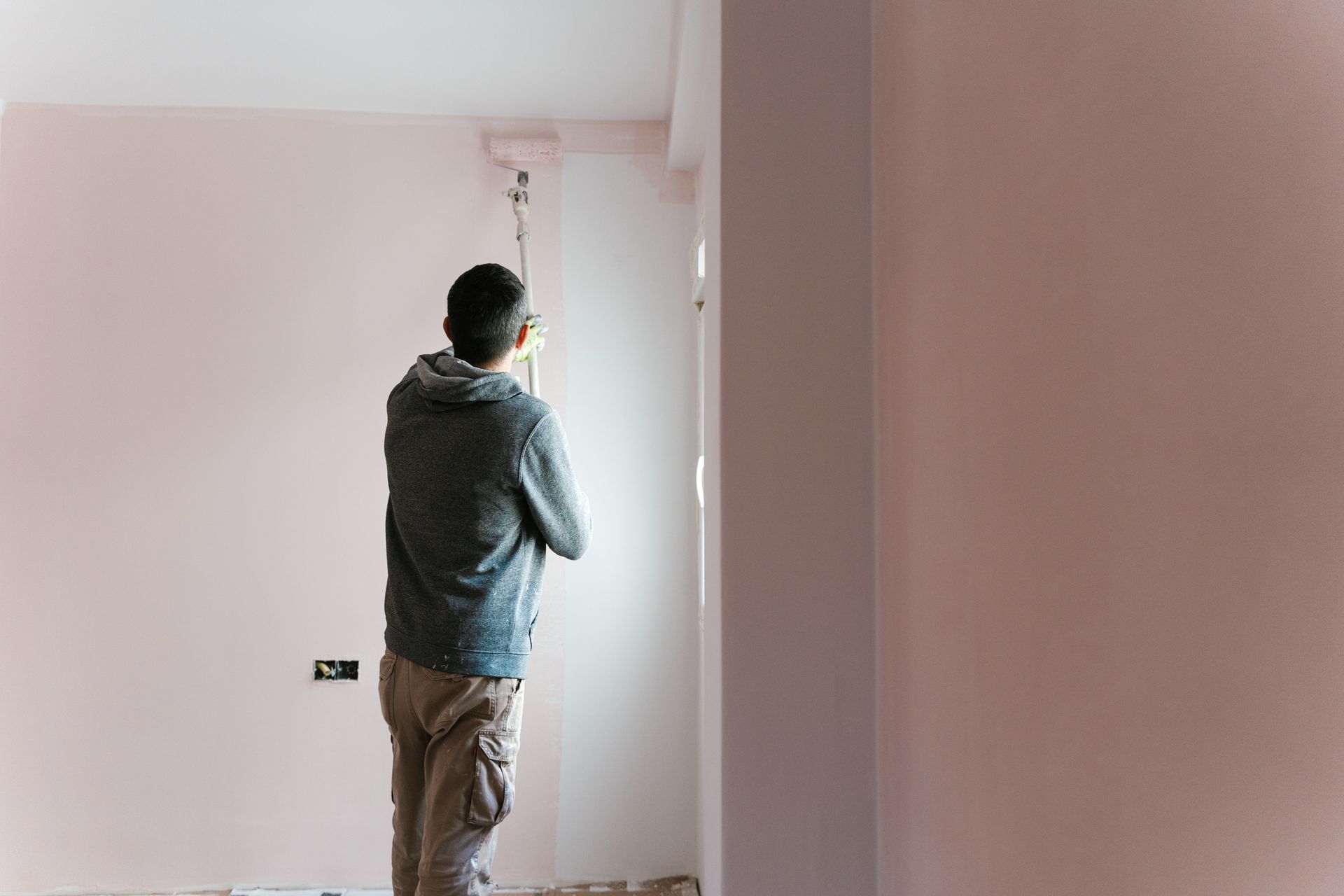 Person standing in a bare room, looking out a window with pink walls and a doorway visible.