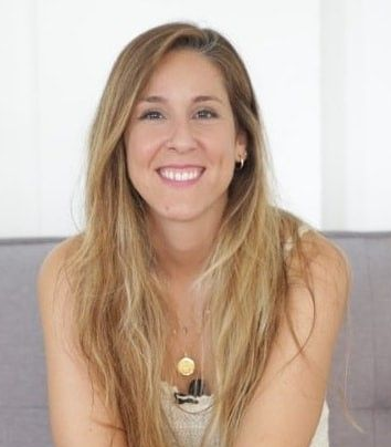 Woman smiling outdoors with brown hair; sunglasses on head; cafe setting.