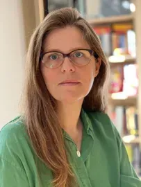 Woman with glasses, long hair, green shirt. Bookshelf in background.
