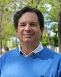 Man in blue sweater, white shirt, smiling outdoors.