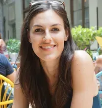 Woman smiling outdoors with brown hair; sunglasses on head; cafe setting.