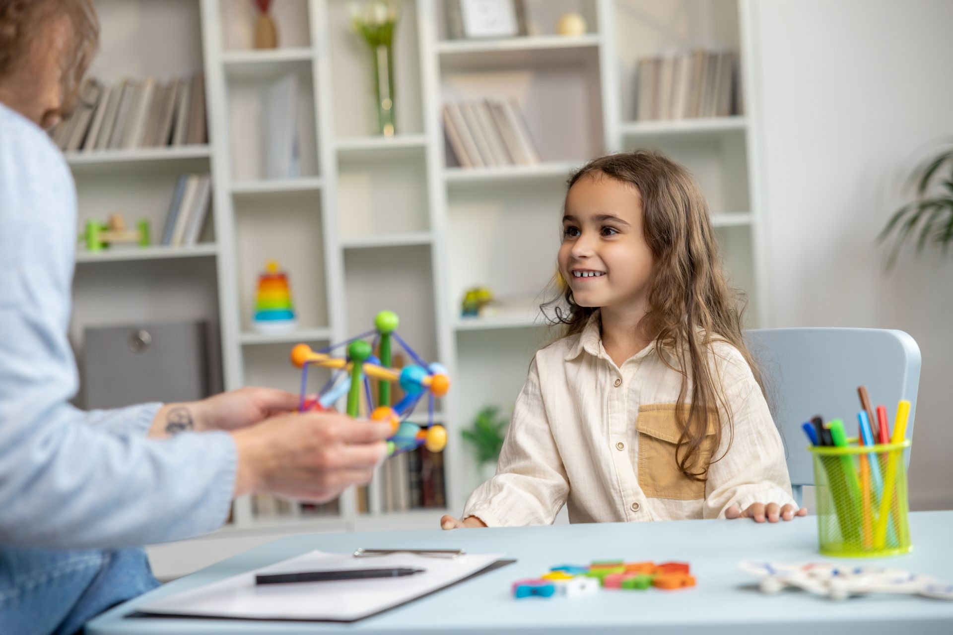 Child smiling at person holding a colorful toy in a therapy room with bookshelves and desk.