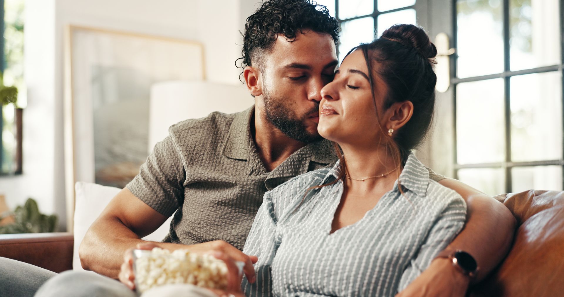 Man kissing woman's cheek on a couch, holding popcorn. They are in a well-lit living room.