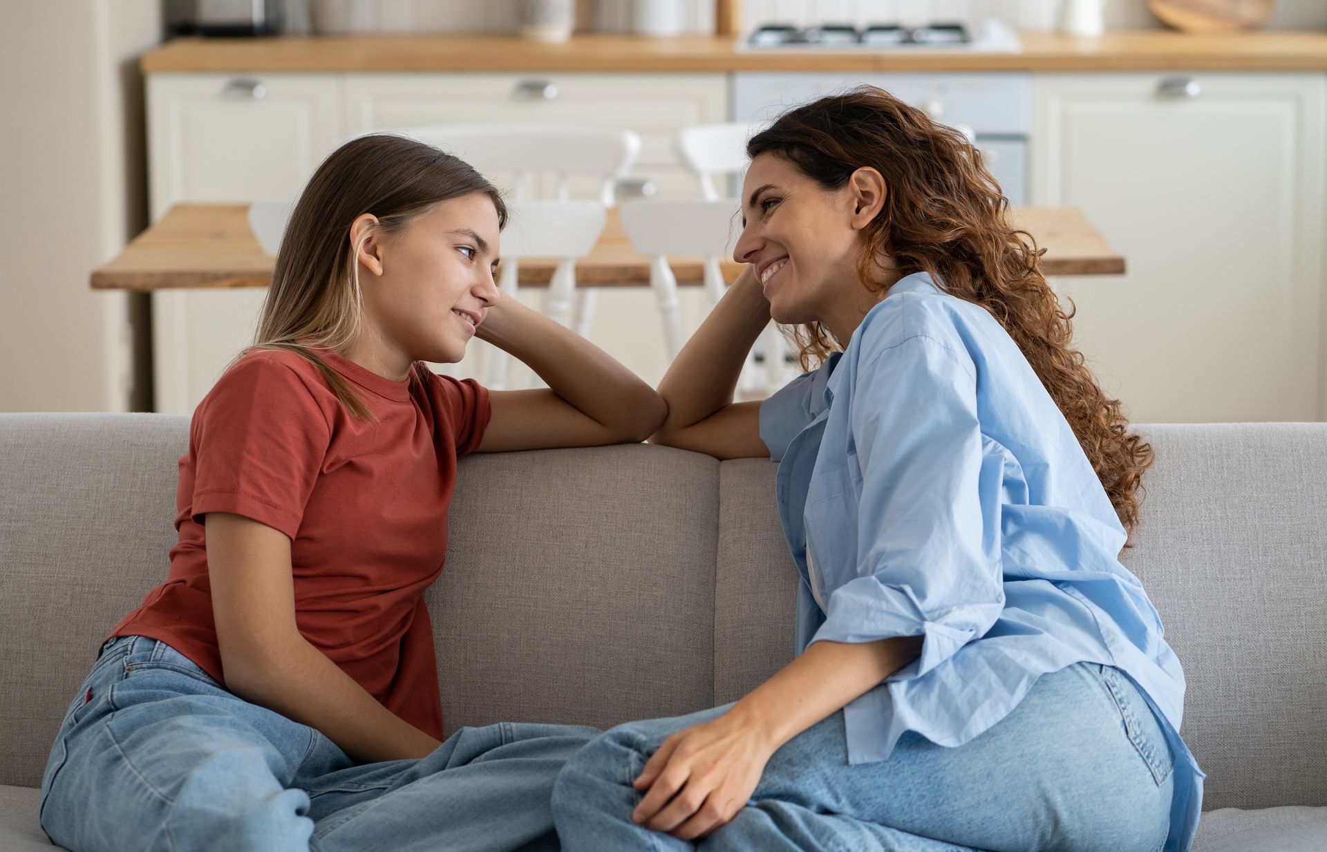 Two people on a couch, looking at each other. One in red shirt and jeans, the other in blue shirt and jeans, smiling.