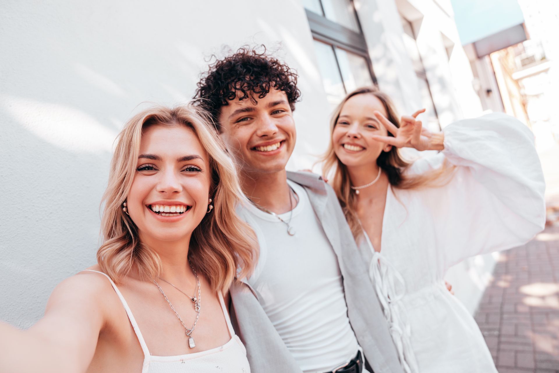 Three smiling people taking a selfie against a white wall. One person makes a peace sign.