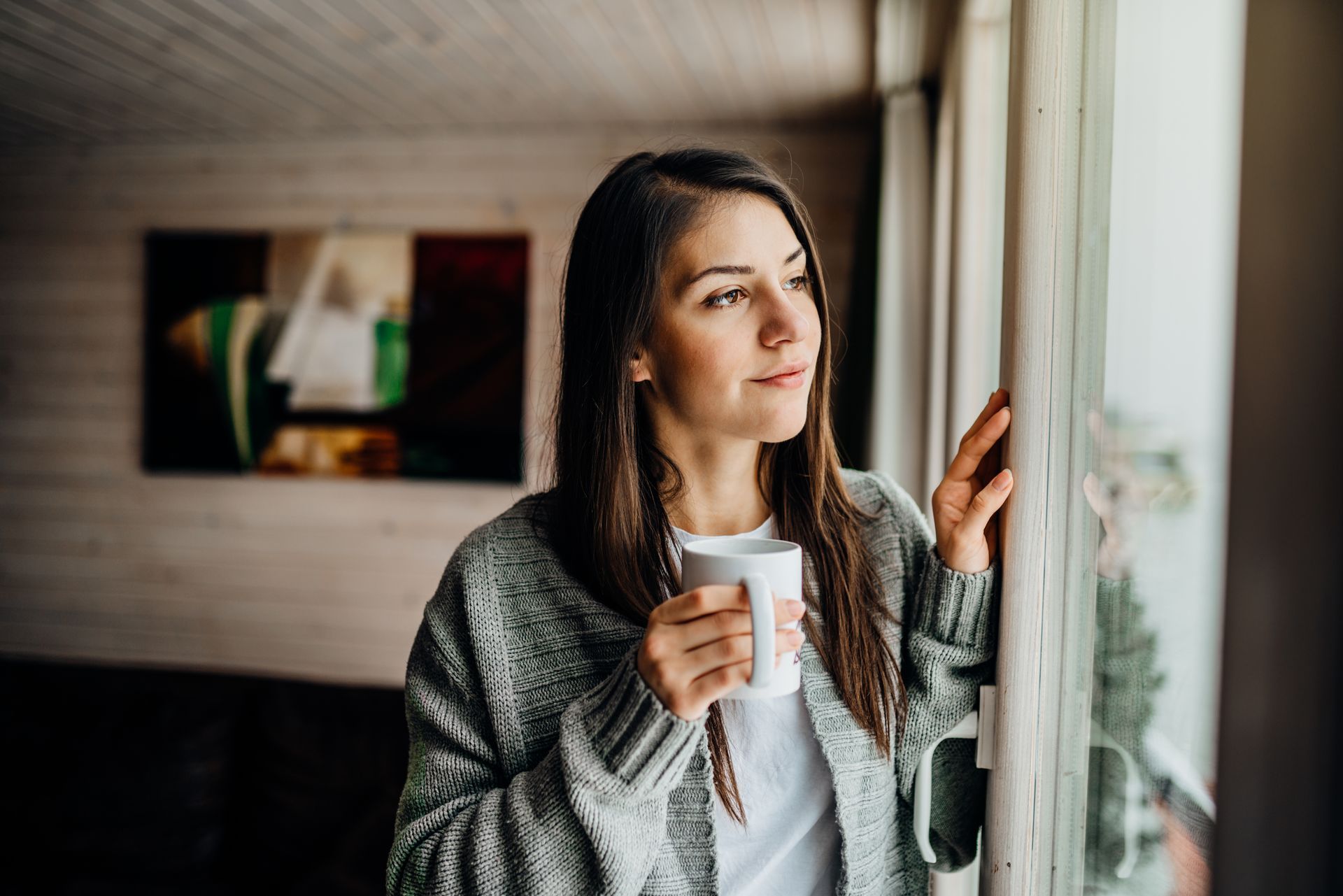 Woman in a gray sweater holding a mug, looking out a window.