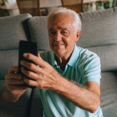 An elderly man is sitting on a couch taking a selfie with his cell phone.