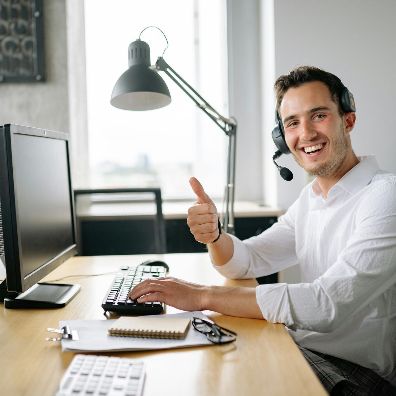 A man is sitting at a desk in front of a computer and giving a thumbs up.