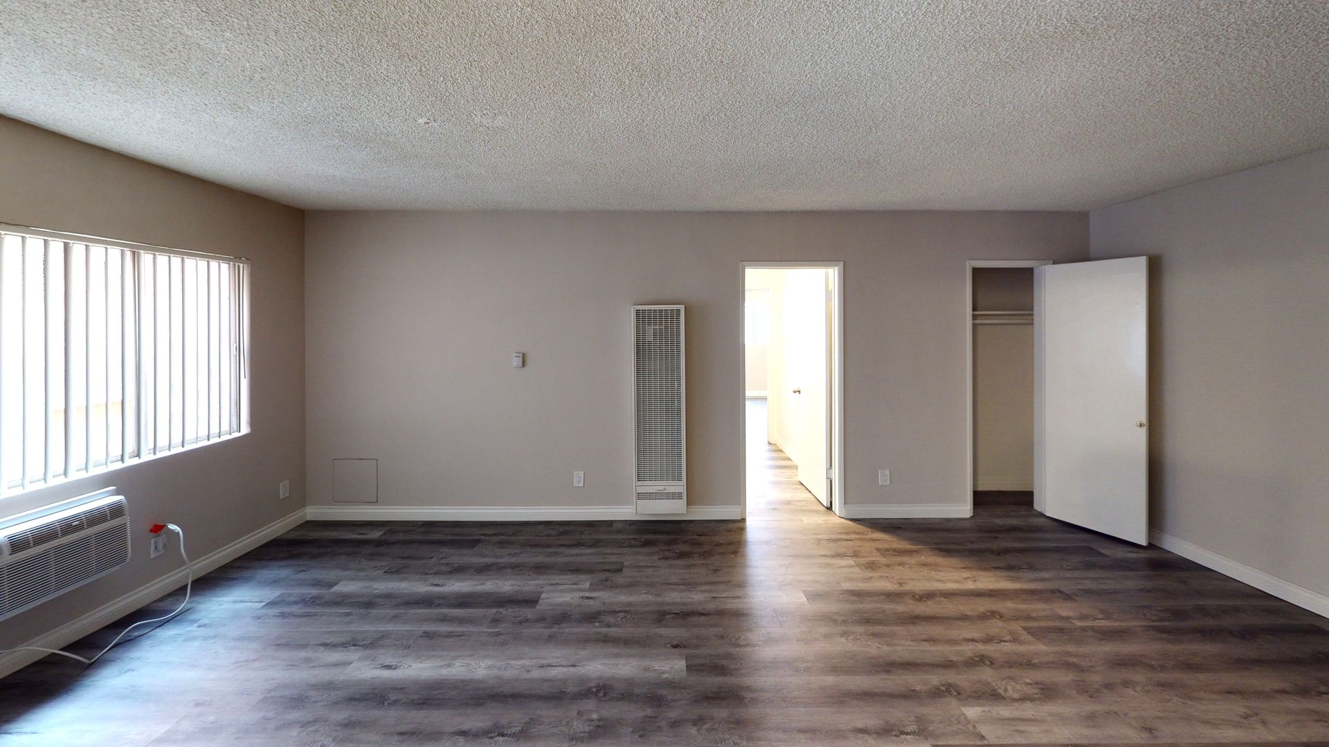 Empty apartment room with wood-look flooring, light gray walls, window with blinds, open doorway and closet.