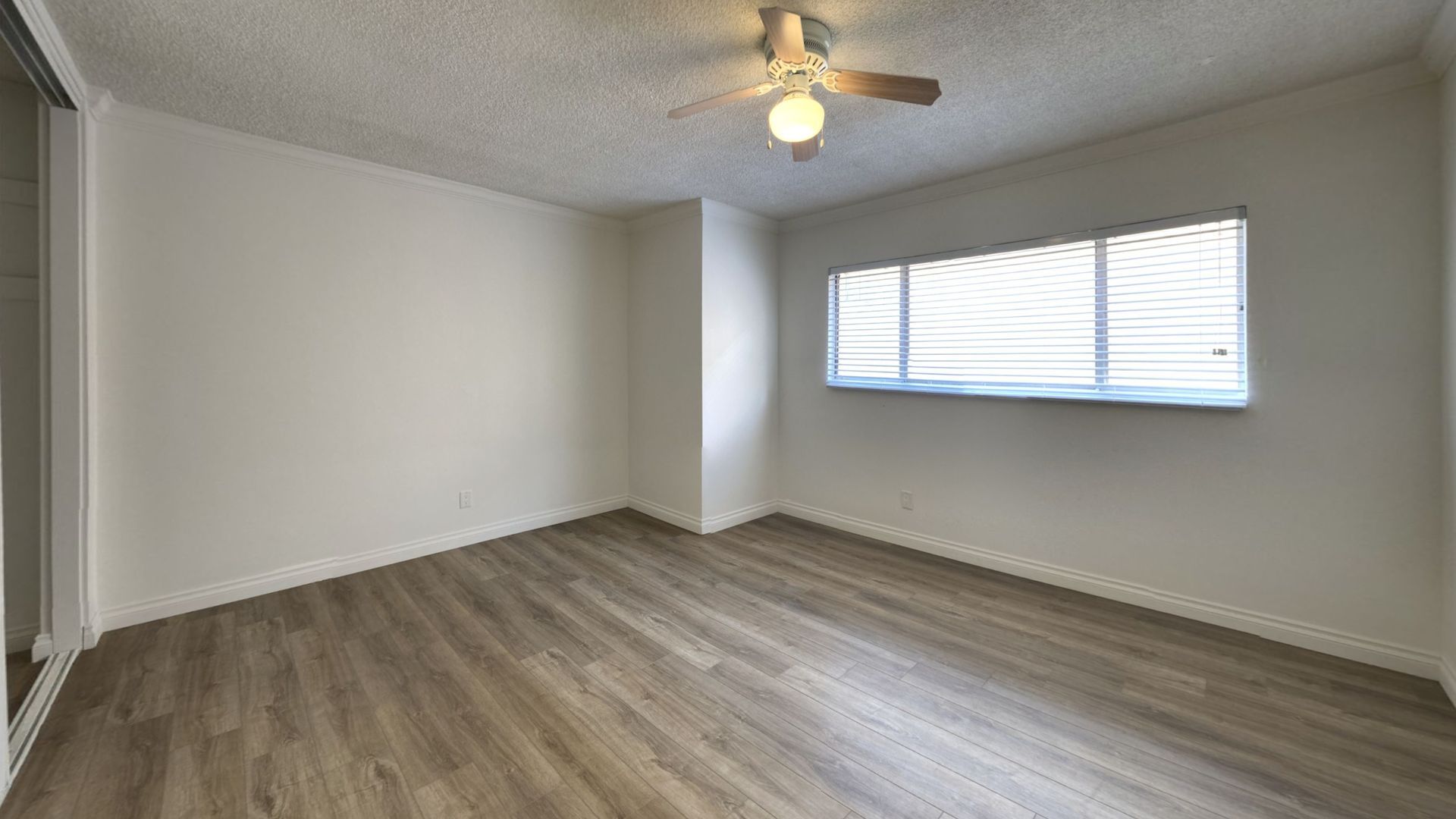 Empty room with light wood floor, white walls, window with blinds, and ceiling fan.
