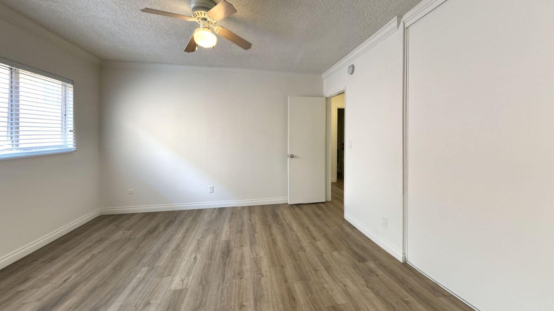 Empty room with wood-look flooring, white walls, a closed door, and a window with blinds. Ceiling fan.