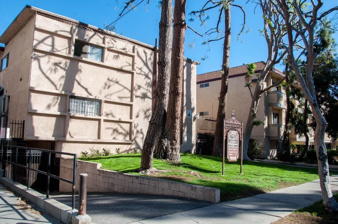 Two-story beige apartment buildings with a small lawn, trees, and a sidewalk.
