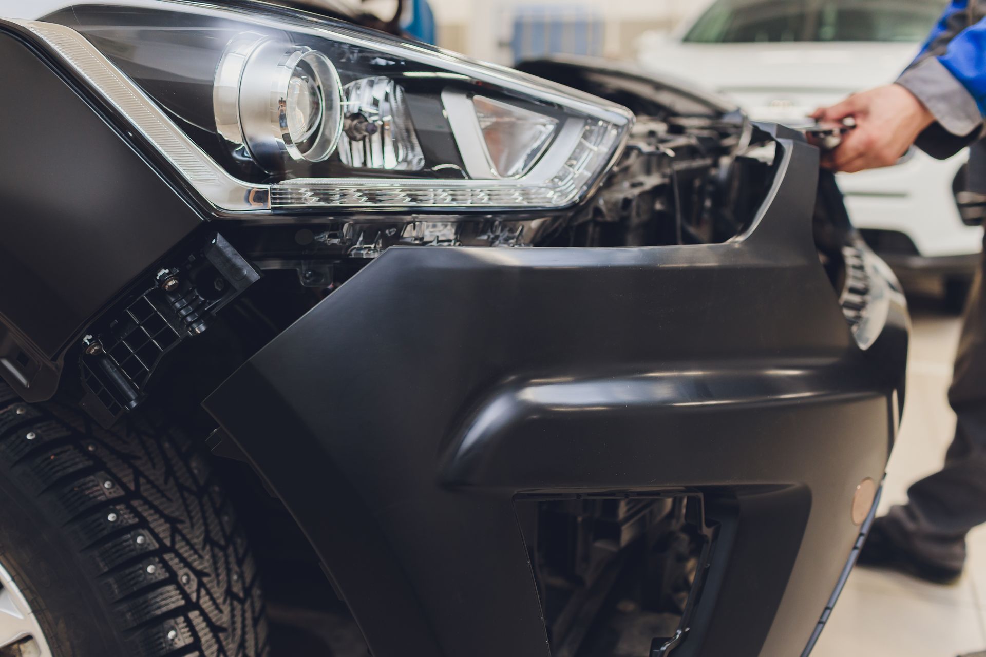 Mechanic working on the front bumper and headlight area of a car in a repair shop.