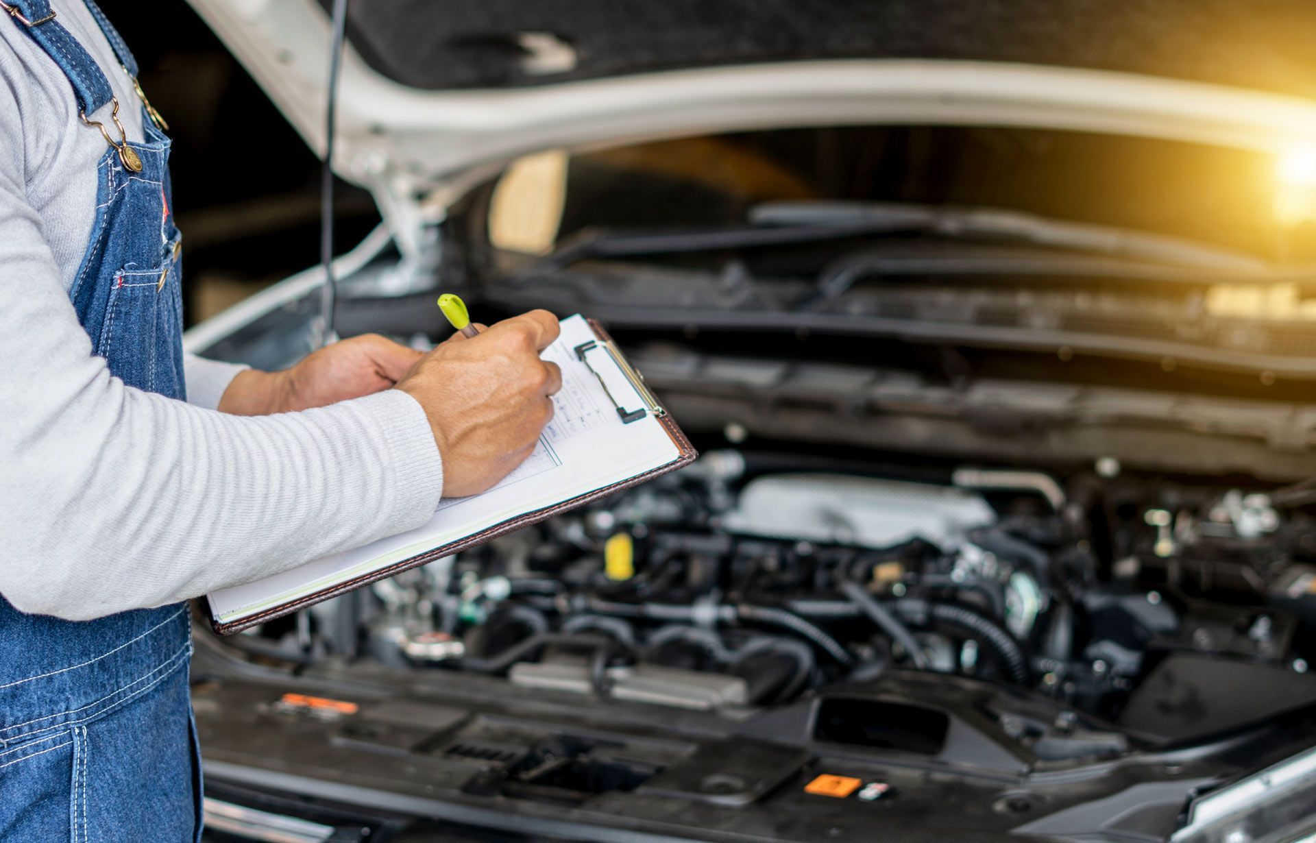 Mechanic inspecting car engine with clipboard inside professional auto body repair shop.