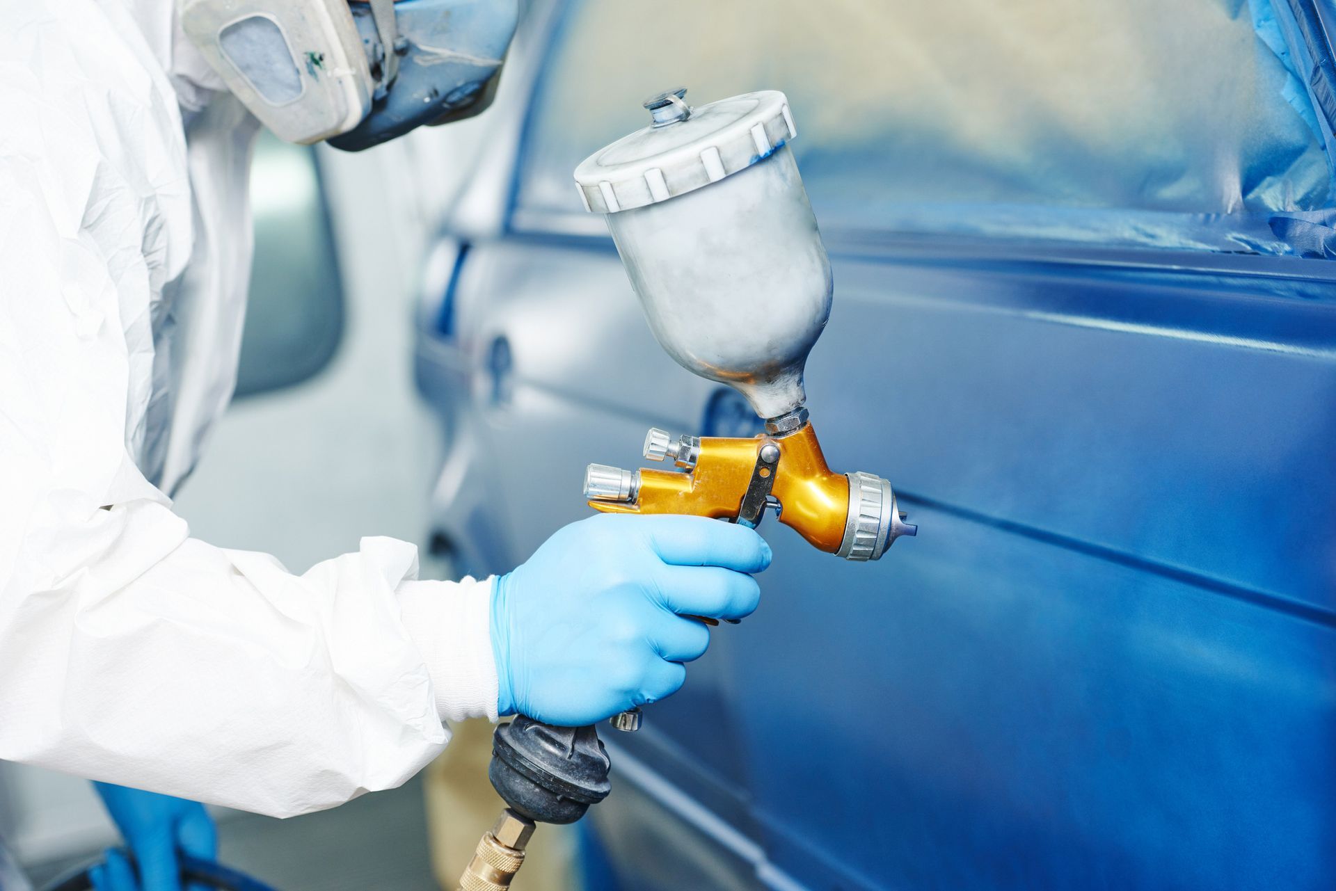 Person in protective gear sprays blue paint on a car in a workshop.