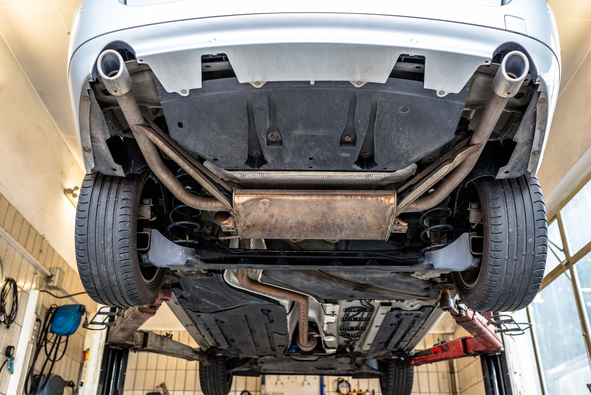 Underside of a car on a lift, showing exhaust system, tires, and undercarriage panels in a garage.