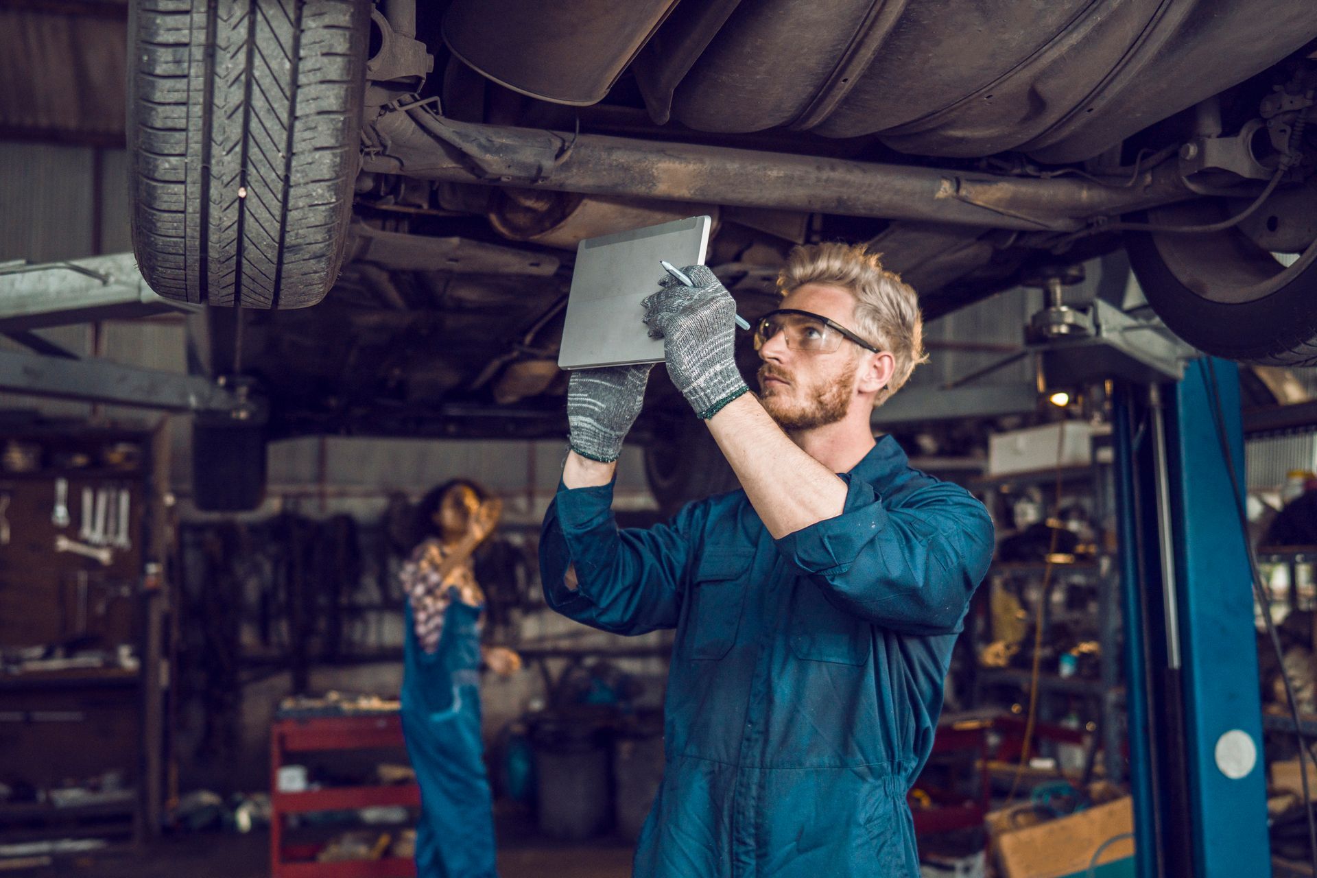Mechanic in blue coveralls examines car underside with a tablet in a garage. Another person is in the background.
