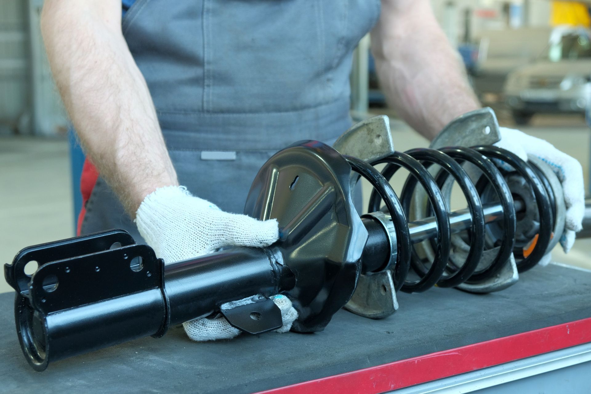 Mechanic holding a black car shock absorber with a coil spring, wearing gloves.