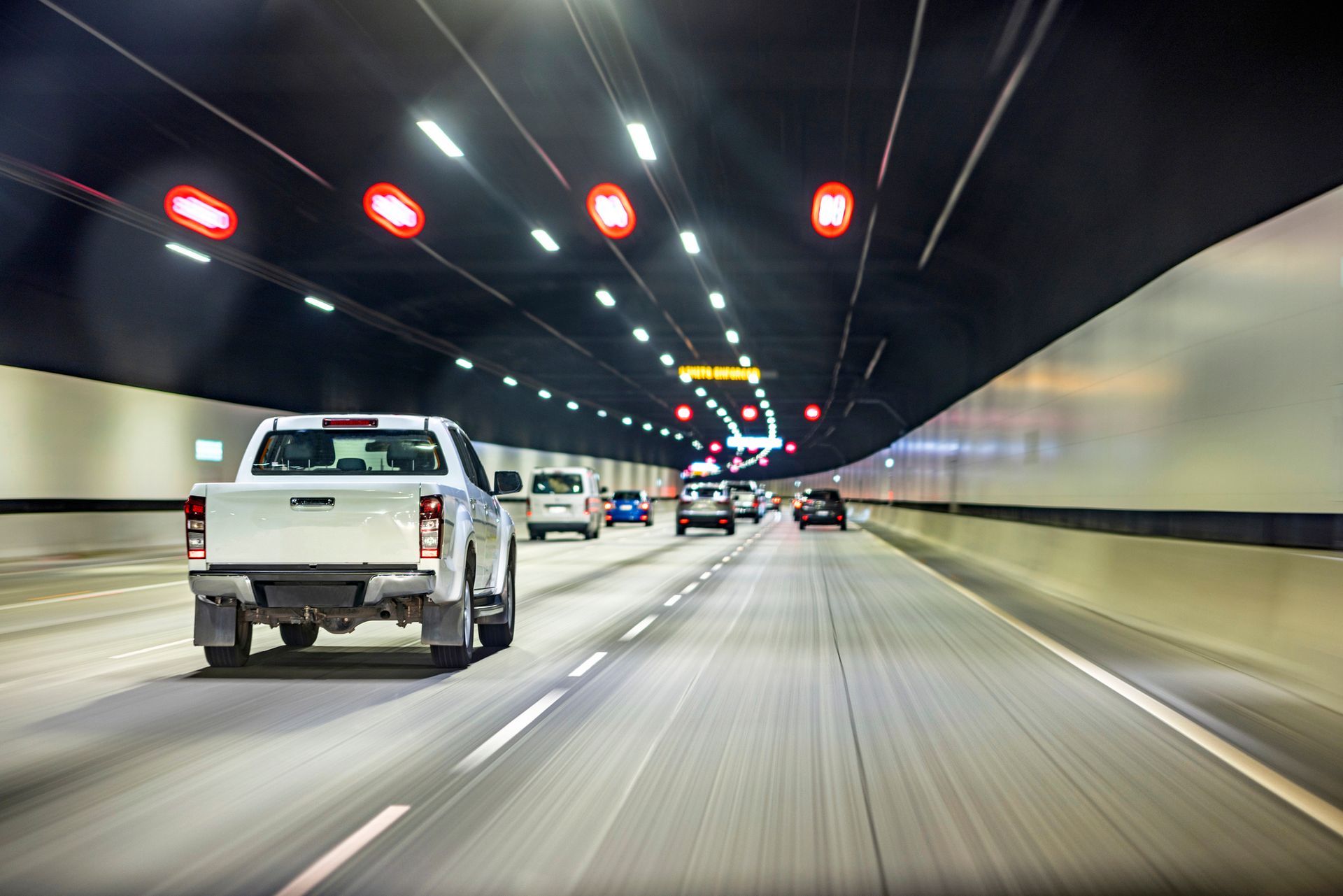 White pickup truck driving in a tunnel with other vehicles. Fluorescent lights and red signals on the ceiling.