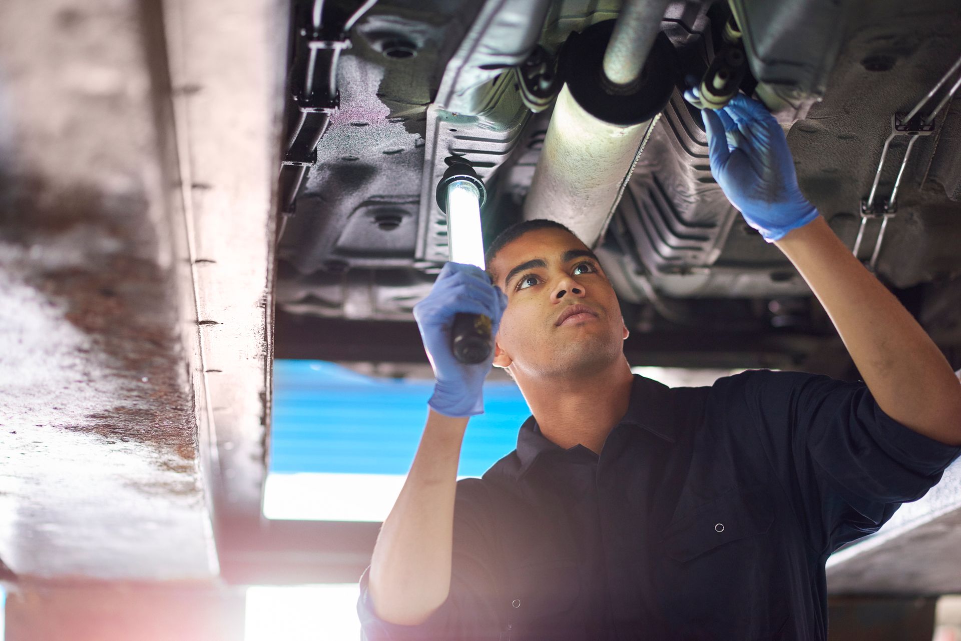 Mechanic inspecting a car's undercarriage with a flashlight, wearing blue gloves.