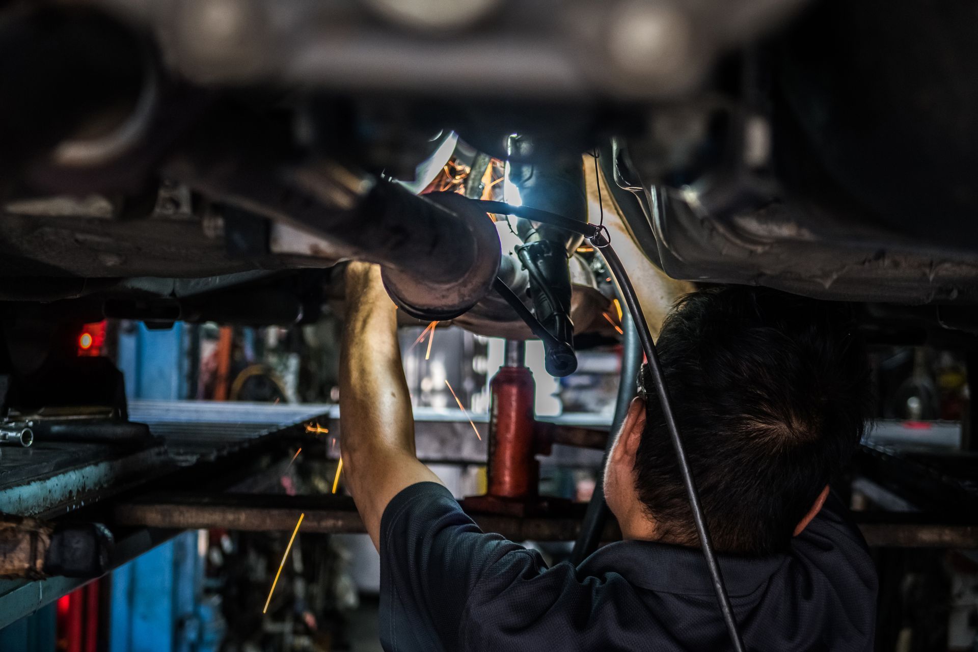 Mechanic welding a car exhaust pipe underneath a vehicle in a shop, sparks flying.