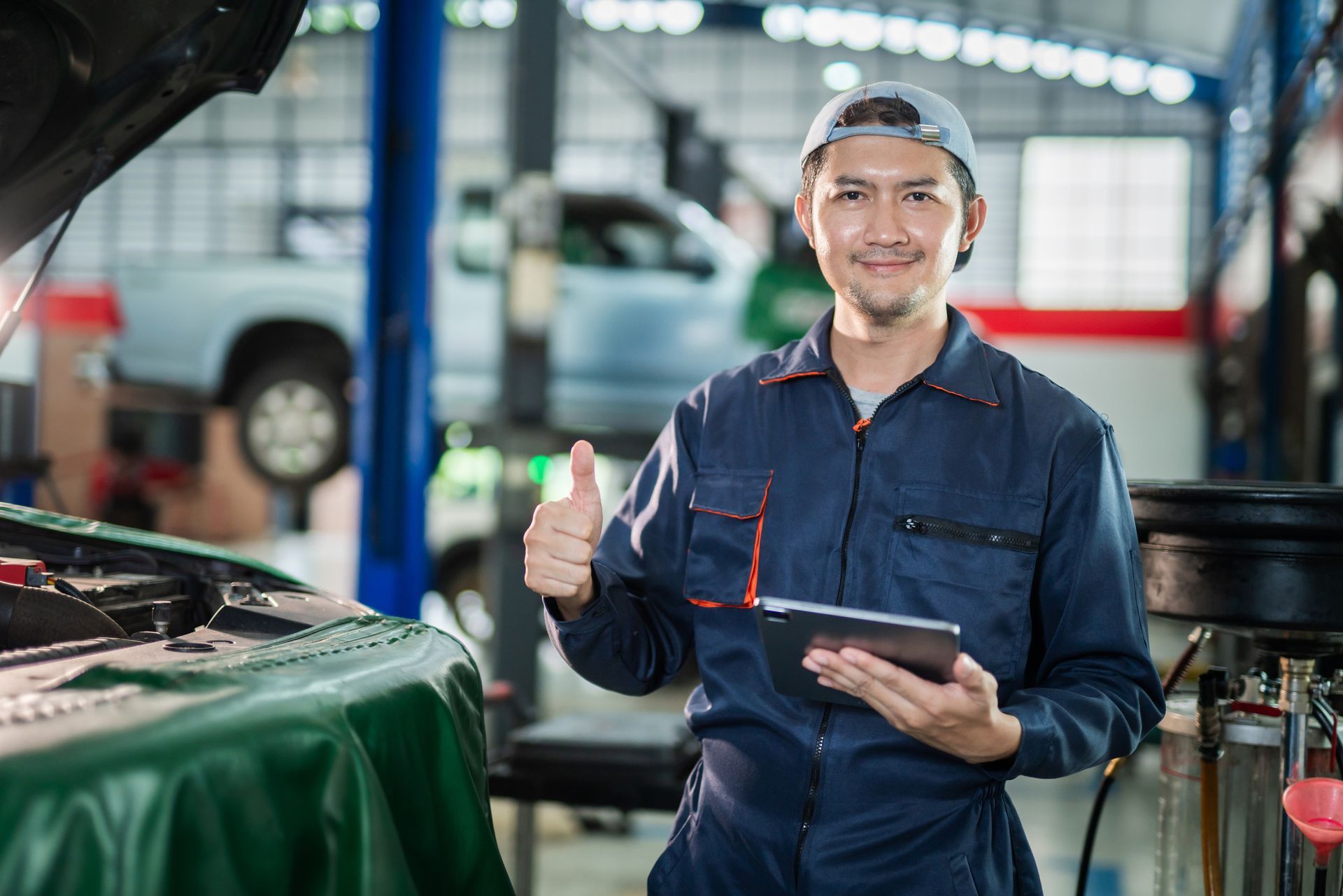Mechanic in blue jumpsuit, holding tablet, gives thumbs-up in auto repair shop.