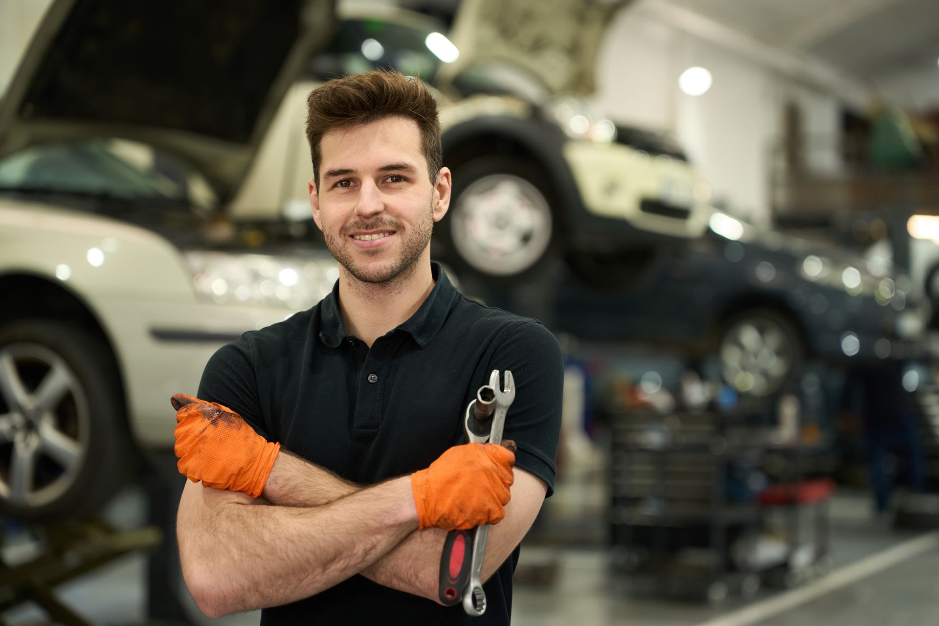 Mechanic in black shirt, orange gloves, holding wrench, arms crossed, smiling in garage with cars.