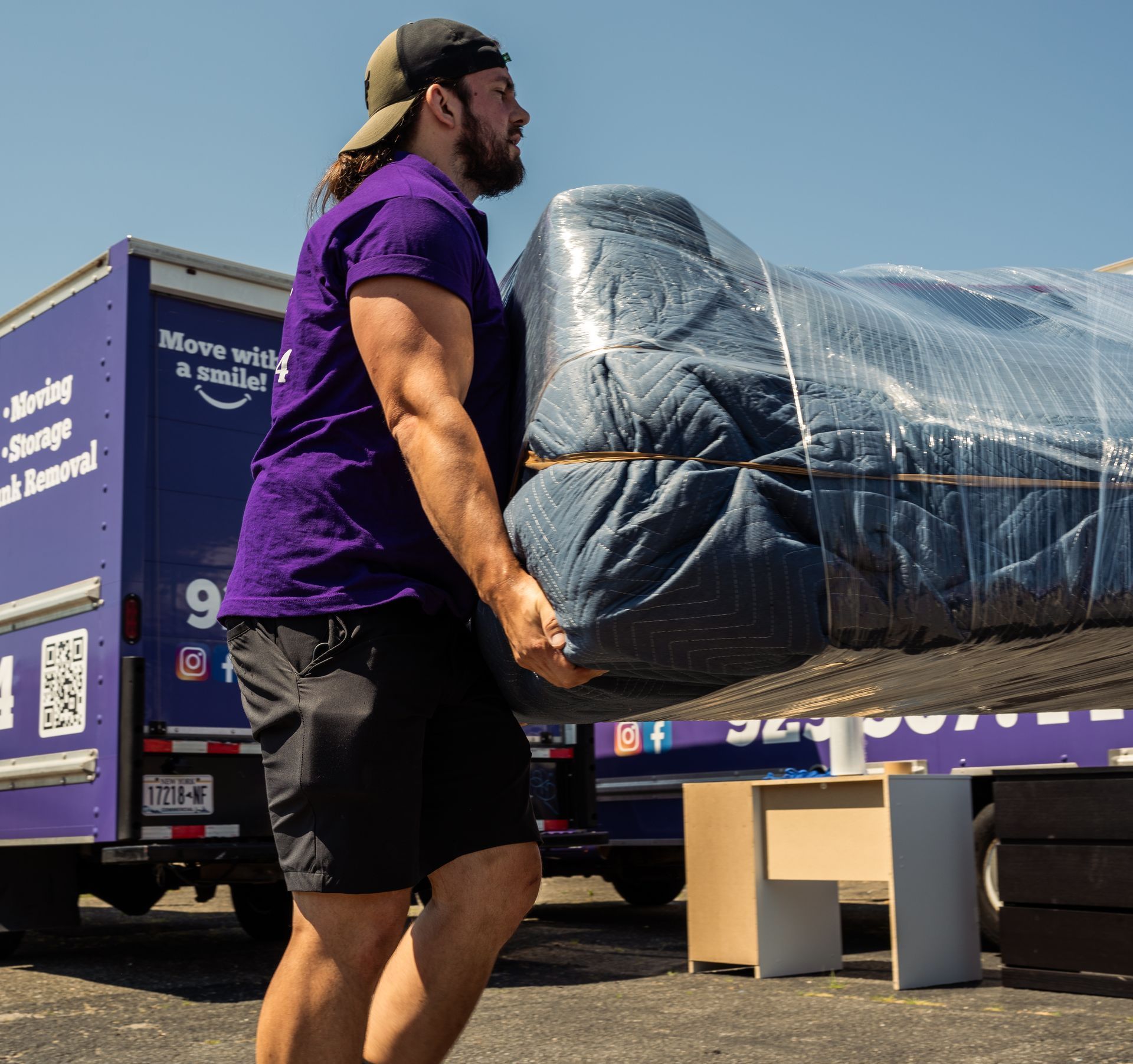 Man in purple shirt carrying a wrapped mattress toward a moving truck.