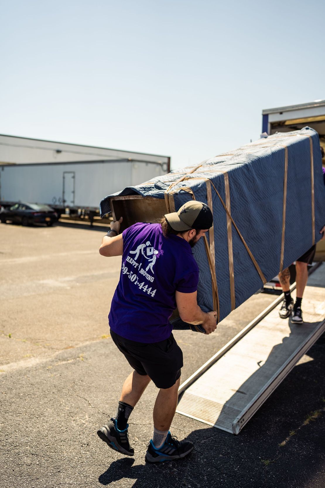 Movers loading a large, wrapped piece of furniture onto a truck ramp on a sunny day.