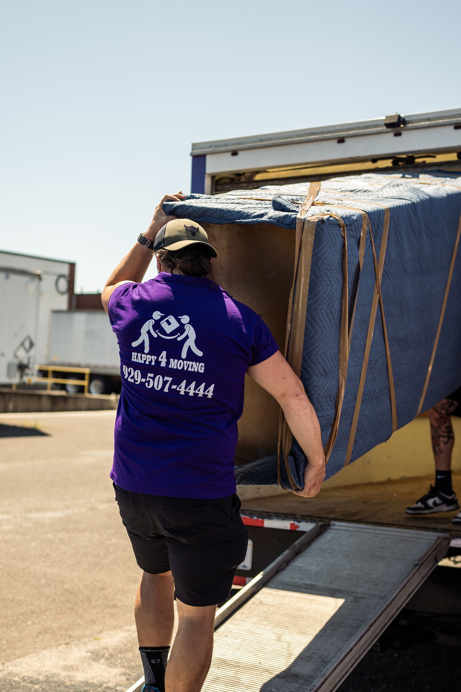 A mover in a purple shirt, wearing a cap, carries a wrapped box into a truck on a ramp.