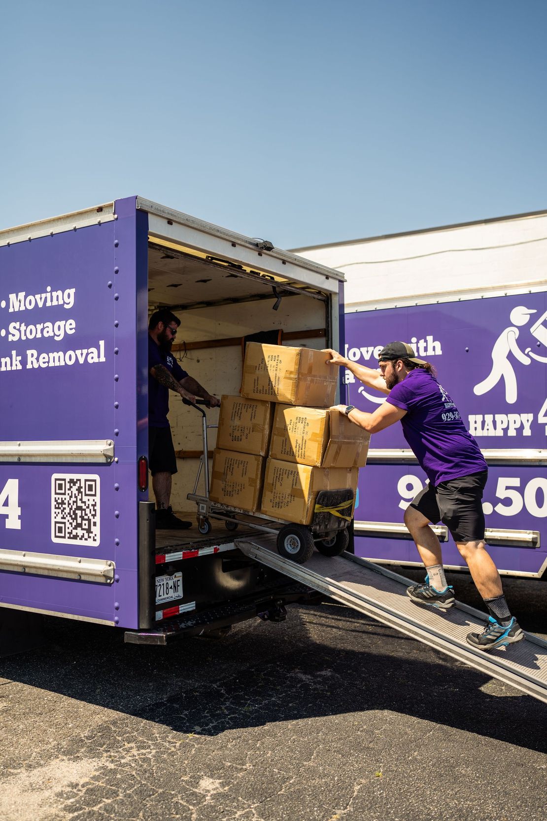 Two men loading boxes into a purple moving truck on a sunny day.