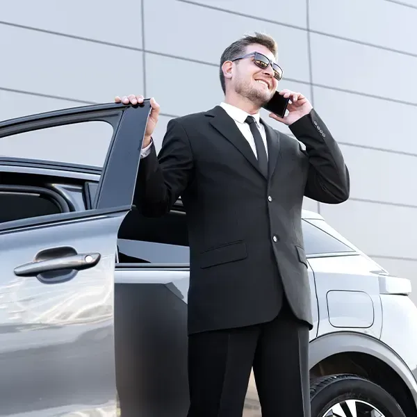 A man in a black suit and sunglasses stands next to a car while talking on his phone, smiling.