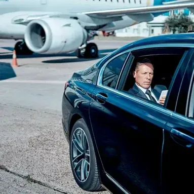 A man in a suit looks out from a black car parked next to a plane on the tarmac. He's holding a phone.