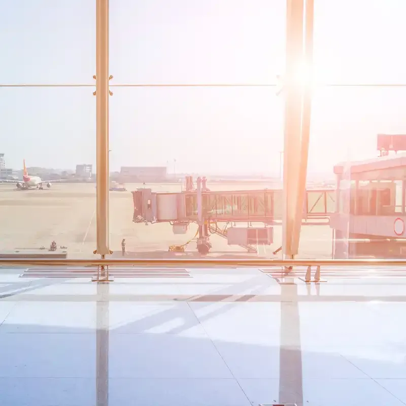 Airport terminal view through large windows, with sunlight, a plane, and a jet bridge.
