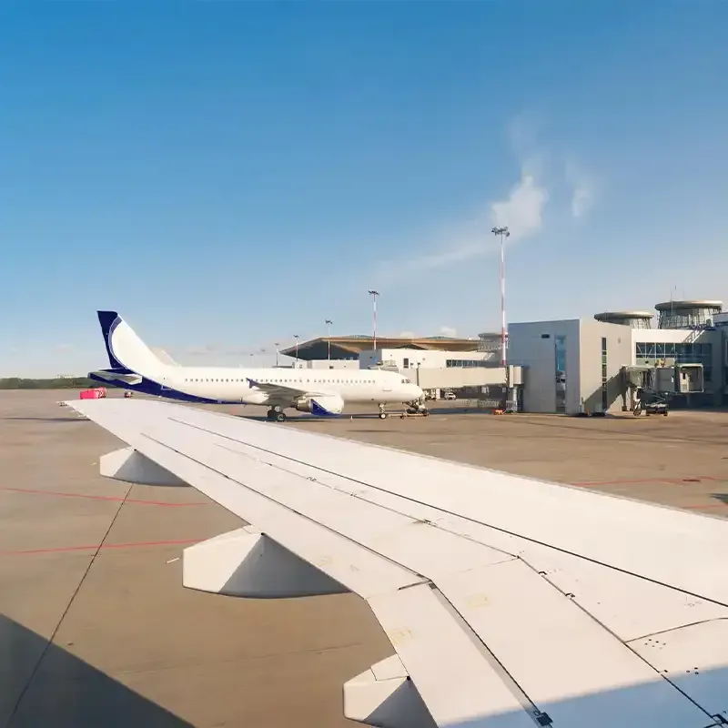 View from a plane window: wing in foreground, airplane and airport terminal in the background under a blue sky.