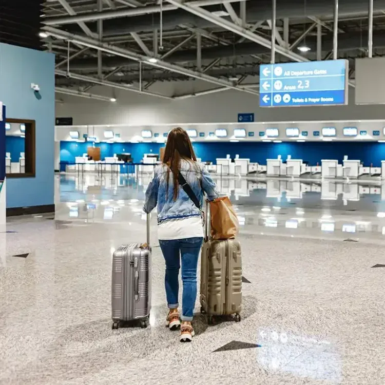 Woman walking through an airport terminal, pulling two suitcases. The terminal has blue accents and departure signs.