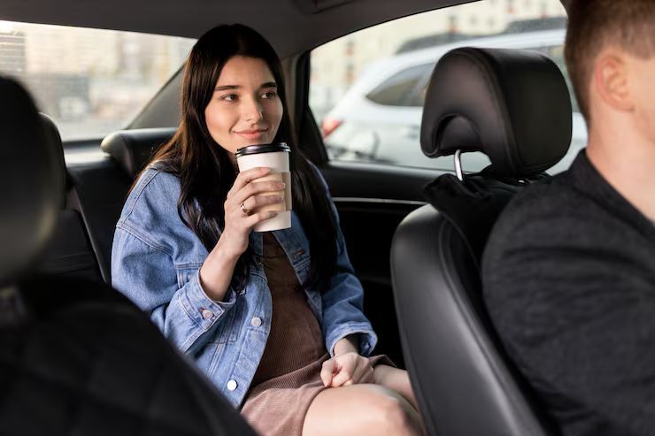 Woman in denim jacket drinks from a coffee cup in the backseat of a car, smiling at the driver.