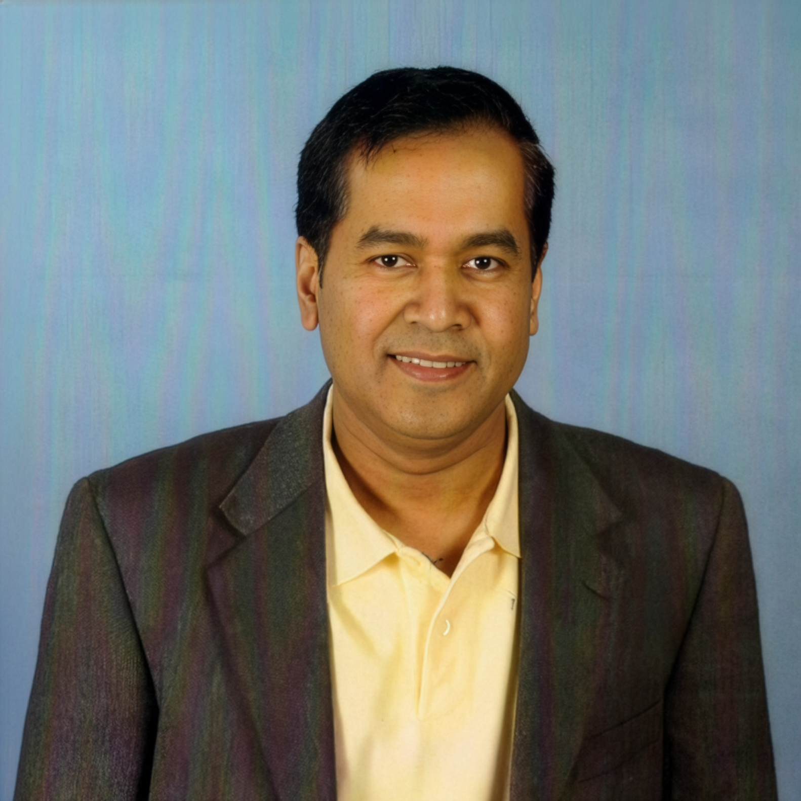 A person in a yellow polo shirt and dark blazer smiles against a light blue, textured backdrop.