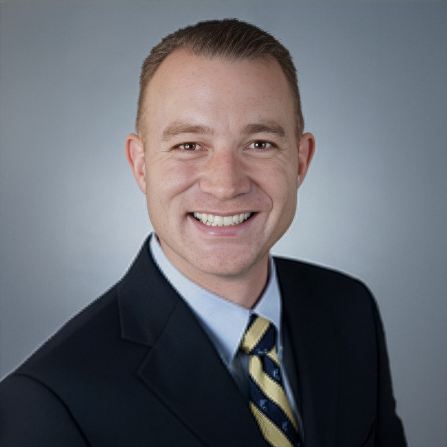 A professional headshot of a smiling person wearing a dark navy suit, light blue dress shirt, and a yellow and blue tie.