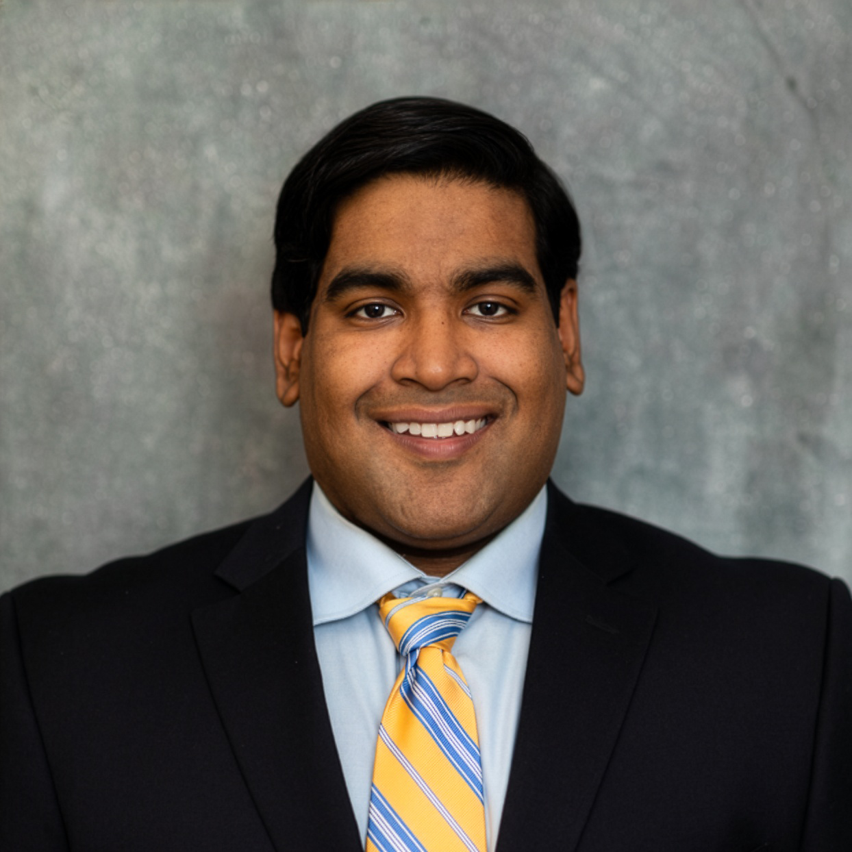 A smiling man in a navy suit, light blue dress shirt, and yellow and blue striped tie against a mottled gray background.
