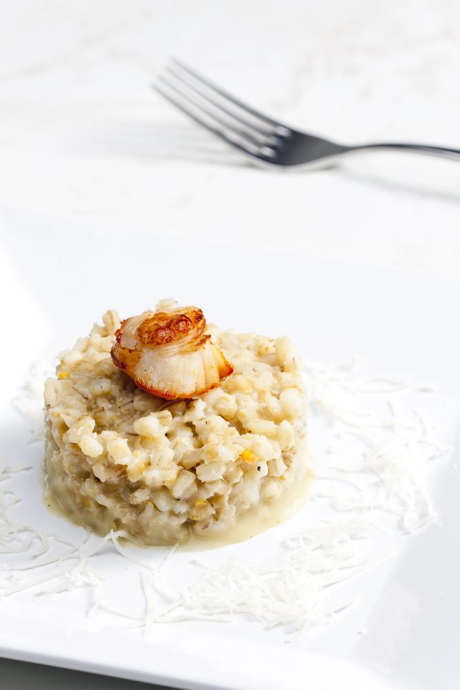 Una capasanta scottata al centro, adagiata su un letto di cremoso risotto su un piatto bianco, con una forchetta sullo sfondo.
