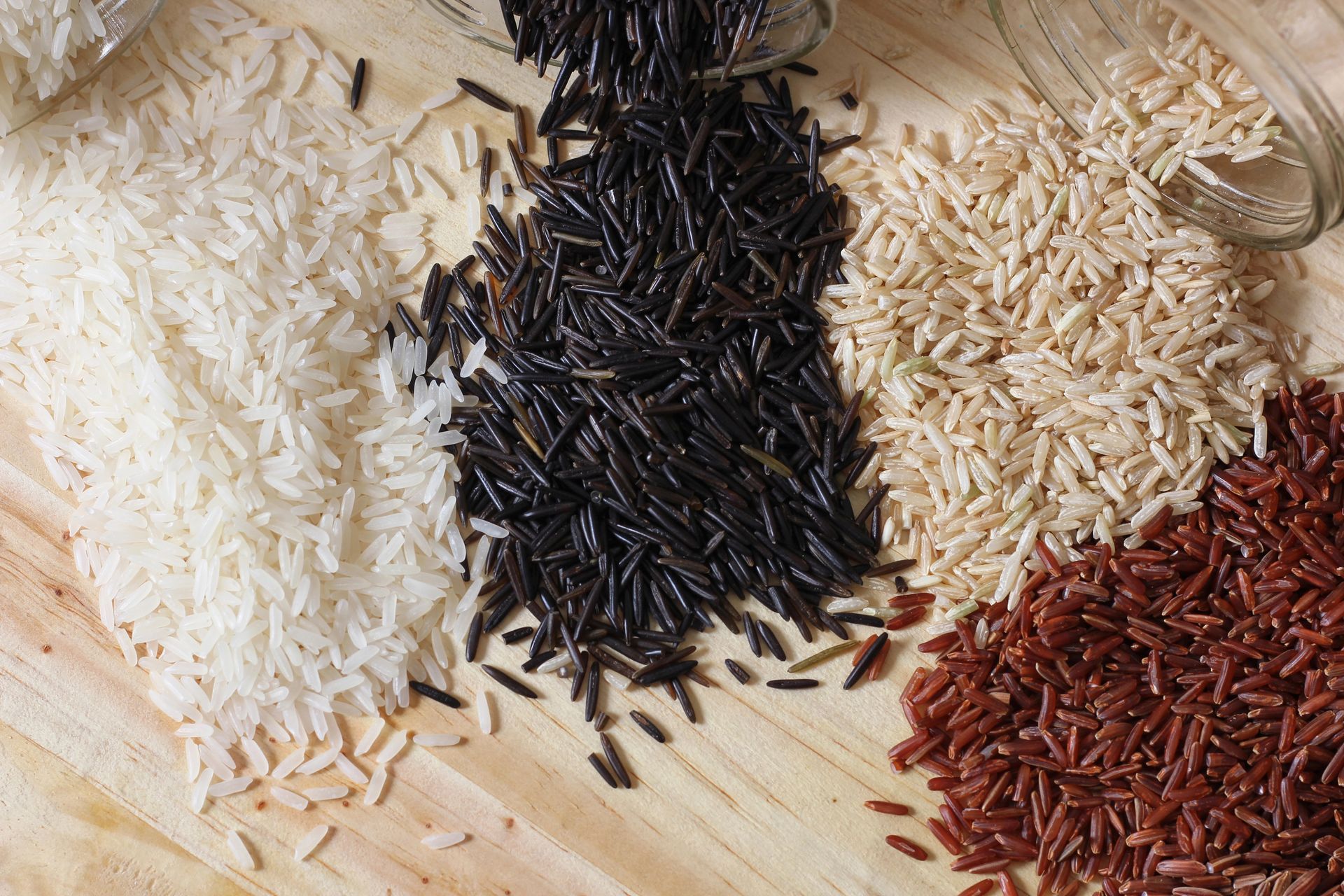 Quattro mucchietti di riso crudo di diverso tipo (bianco, nero, integrale e rosso) su una superficie di legno chiaro.