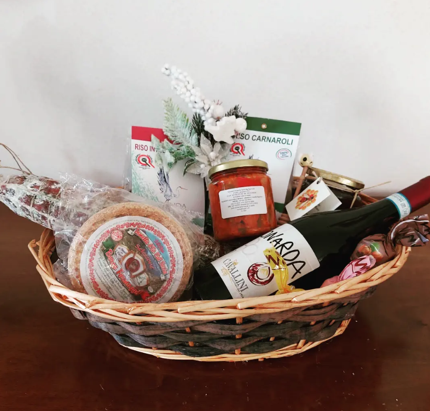 Un cesto regalo intrecciato contenente una bottiglia di vino rosso, un barattolo di salsa, prodotti in scatola e snack confezionati, adagiato su un tavolo di legno.