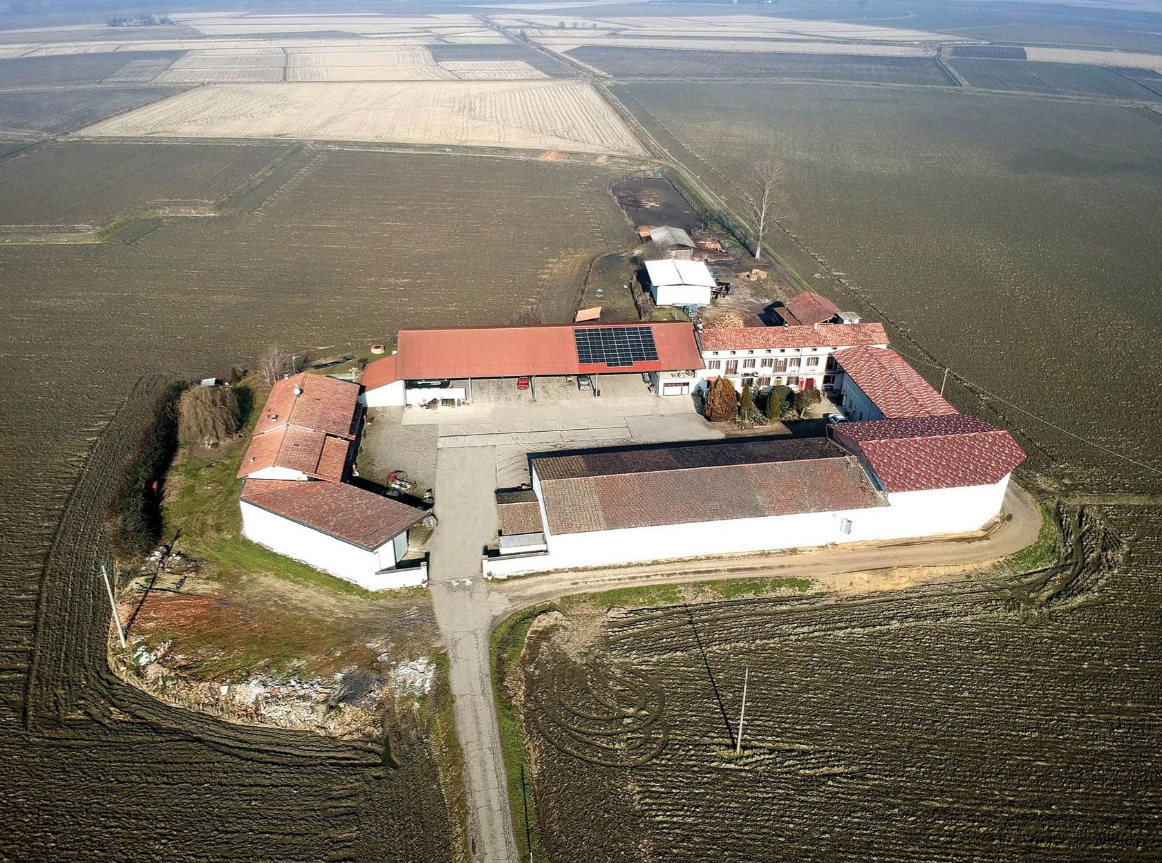 Una vista aerea mostra due trattori su una strada sterrata rurale tra campi agricoli arati.