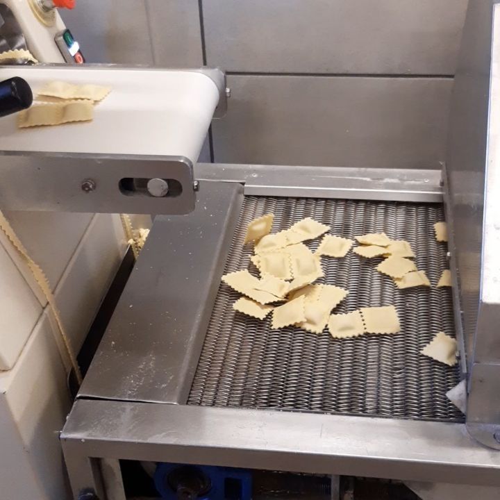 I ravioli scorrono su un nastro trasportatore e cadono su una griglia metallica all'interno di una macchina industriale per la lavorazione della pasta.