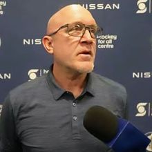 Man wearing glasses speaks into a microphone; Nissan and logo backdrop.