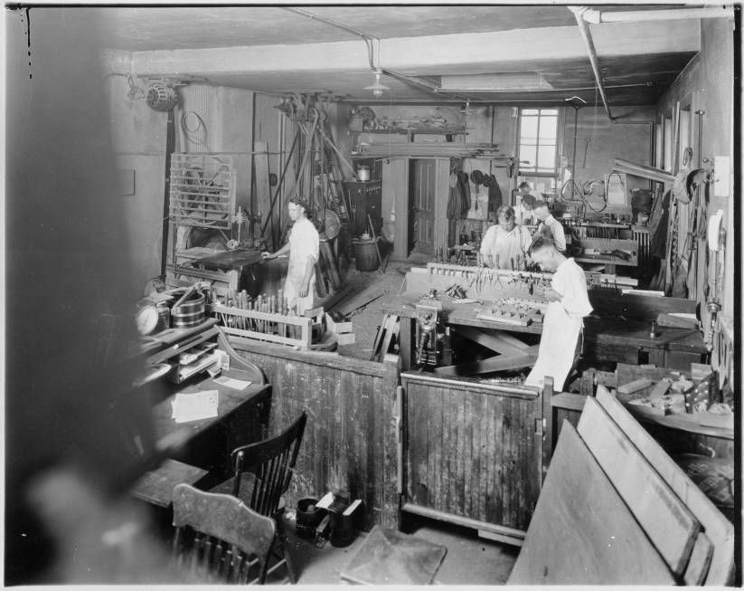 Busy workshop with several people working at tables amid tools, wood, and supplies